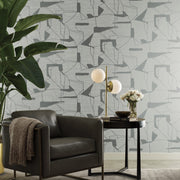 Living Room - A modern with a gray armchair, a side table holding a double-bulb lamp, and a vase of white flowers. The backdrop is Fog & Silver Abstract Geo Wallpaper by York Wallcoverings from the Modern Metals Collection in abstract patterns, complemented by a large potted plant