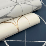 Two rolls from York Wallcoverings—one Fog & Silver Sun Circles Wallpaper in light cream with metallic silver geometric lines and another in white with gold accents—rest on a dark blue surface adorned with white geometric patterns.