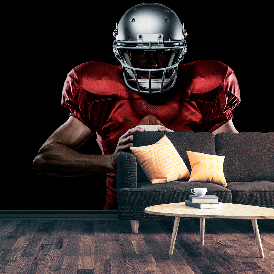A Decor2Go Wallpaper Mural featuring a football player in full gear crouches behind a dark sofa, gripping a football and staring intently forward, against a dark background with a wooden floor. This scene perfectly captures the essence of the Football Player Wallpaper Mural.