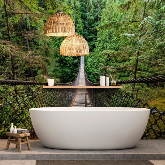A luxurious freestanding bathtub set on a concrete floor with a backdrop of a custom Decor2Go Wallpaper Mural depicting a lush green forest and a wooden suspension bridge. Above the tub hang two woven pendant lights, enhancing