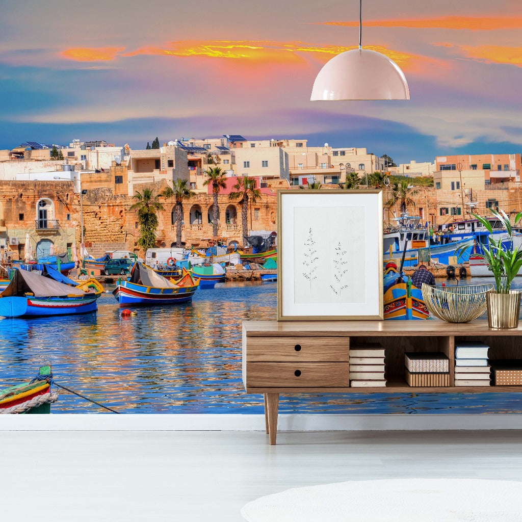 A vibrant living room with a scenic Forever Malta Wallpaper Mural from Decor2Go Wallpaper Mural of a Mediterranean coastline featuring colorful boats and historic architecture. A modern sideboard and lamp complete the decor.