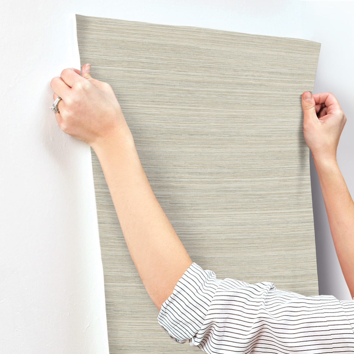 A person in a striped shirt is applying a piece of York Wallcoverings Fountain Grass Camel Wallpaper Brown (60 Sq.Ft.) with a natural aesthetic to a white wall. Holding the wallpaper at both corners, they ensure it is properly aligned for easy installation.