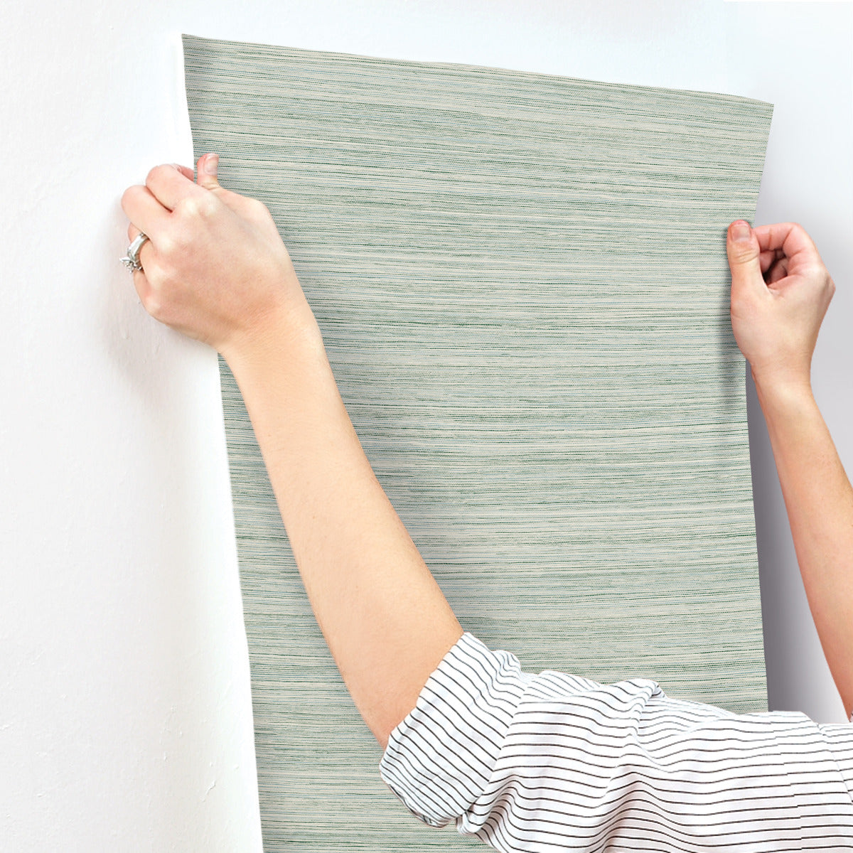 A person with rolled-up sleeves is applying a strip of York Wallcoverings Fountain Grass Smokey Blue Wallpaper Blue (60 Sq.Ft.) to a smooth, white wall. Using both hands to align the wallpaper at the top edge, they ensure an easy installation that enhances the room's natural aesthetic.