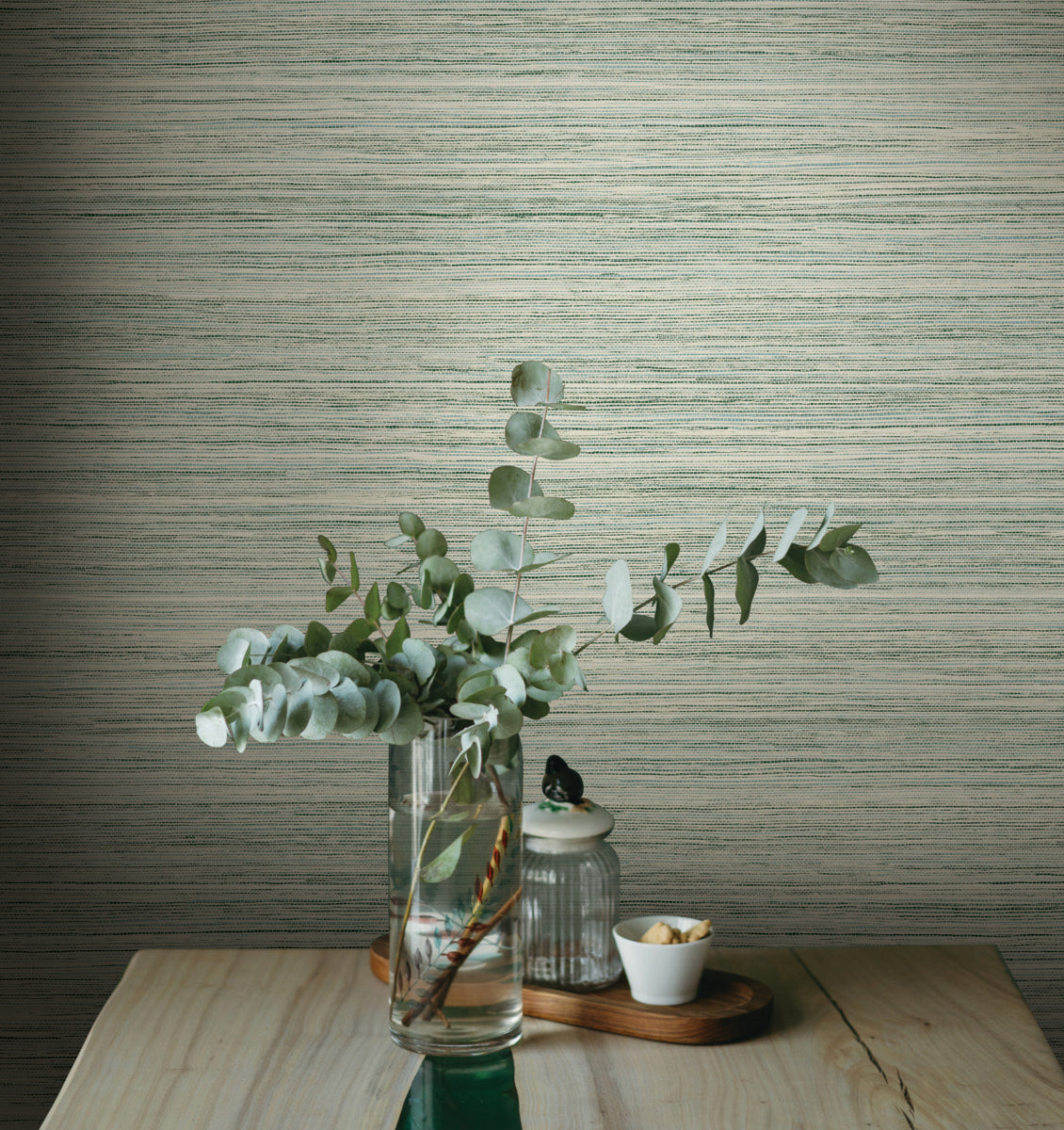 A glass vase holds eucalyptus branches and sits on a light wooden table. Nearby, a small lidded jar and a tiny white bowl with nuts are arranged. The backdrop features York Wallcoverings' Fountain Grass Camel Wallpaper Brown (60 Sq.Ft.). The scene evokes a minimalist and natural aesthetic.