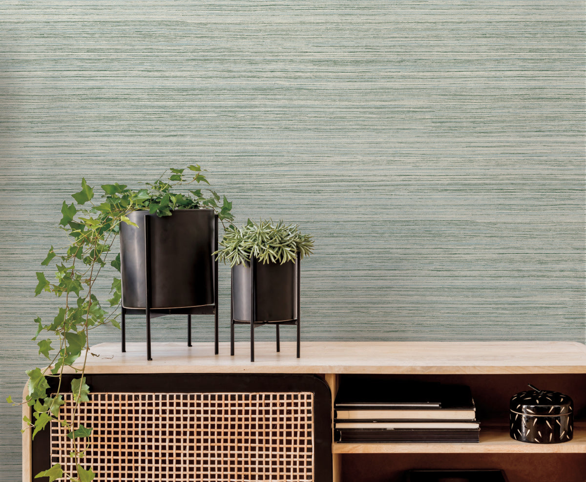 A modern interior features a light wood shelf with a rattan front panel, offering an easy installation and blending seamlessly into the natural aesthetic. On top are two black metal planters of different heights, each holding green plants. The background is a textured, light green York Wallcoverings Fountain Grass Smokey Blue Wallpaper Blue (60 Sq.Ft.), adding a soothing contrast to the setup.