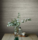 A vase with eucalyptus branches sits on a wooden table against a textured, beige wall adorned with York Wallcoverings Fountain Grass Camel Wallpaper Brown (60 Sq.Ft.). Next to the vase is a small, covered glass jar and a white bowl of nuts, all arranged on a wooden tray, enhancing the natural aesthetic of the space.