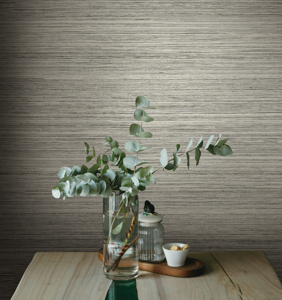 A vase with eucalyptus branches sits on a wooden table against a textured, beige wall adorned with York Wallcoverings Fountain Grass Camel Wallpaper Brown (60 Sq.Ft.). Next to the vase is a small, covered glass jar and a white bowl of nuts, all arranged on a wooden tray, enhancing the natural aesthetic of the space.