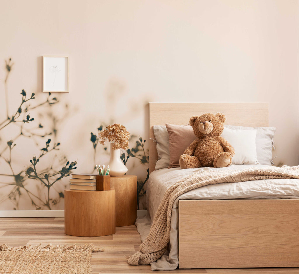 Cozy bedroom featuring a large bed with beige linens and a plush teddy bear sitting on it. Next to the bed is a wooden nightstand with books and a vase of dried flowers, under Decor2Go Wallpaper Mural's Fragile Flowers Wallpaper Mural.