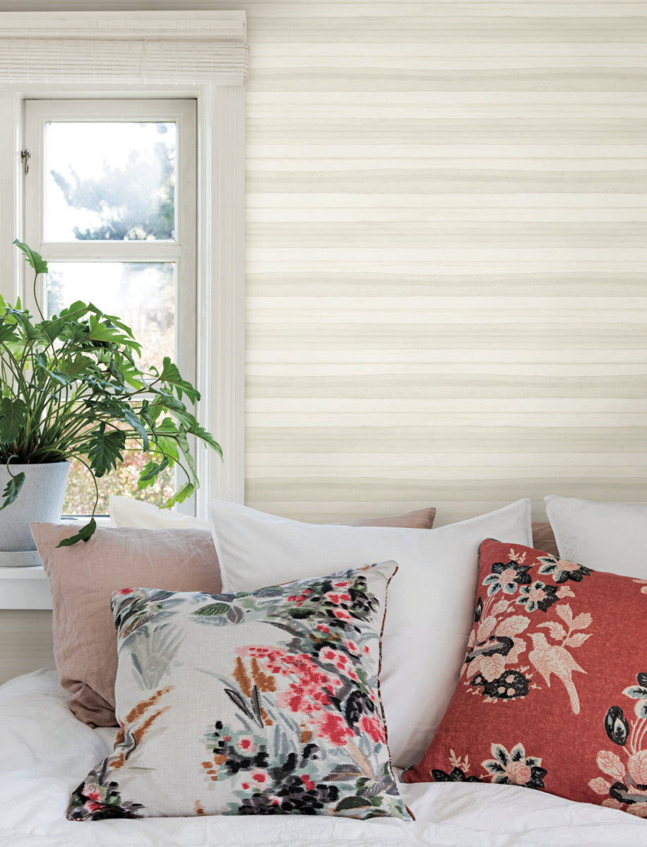 A cozy interior scene featuring a bed with white linens, accented by decorative pillows with floral patterns. A potted plant with tropical foliage sits on the windowsill, and soft, natural light filters in through the window. The walls are adorned with Pandora Leaf Cotton Wallpaper White/Off White (60 Sq.Ft.) by York Wallcoverings.