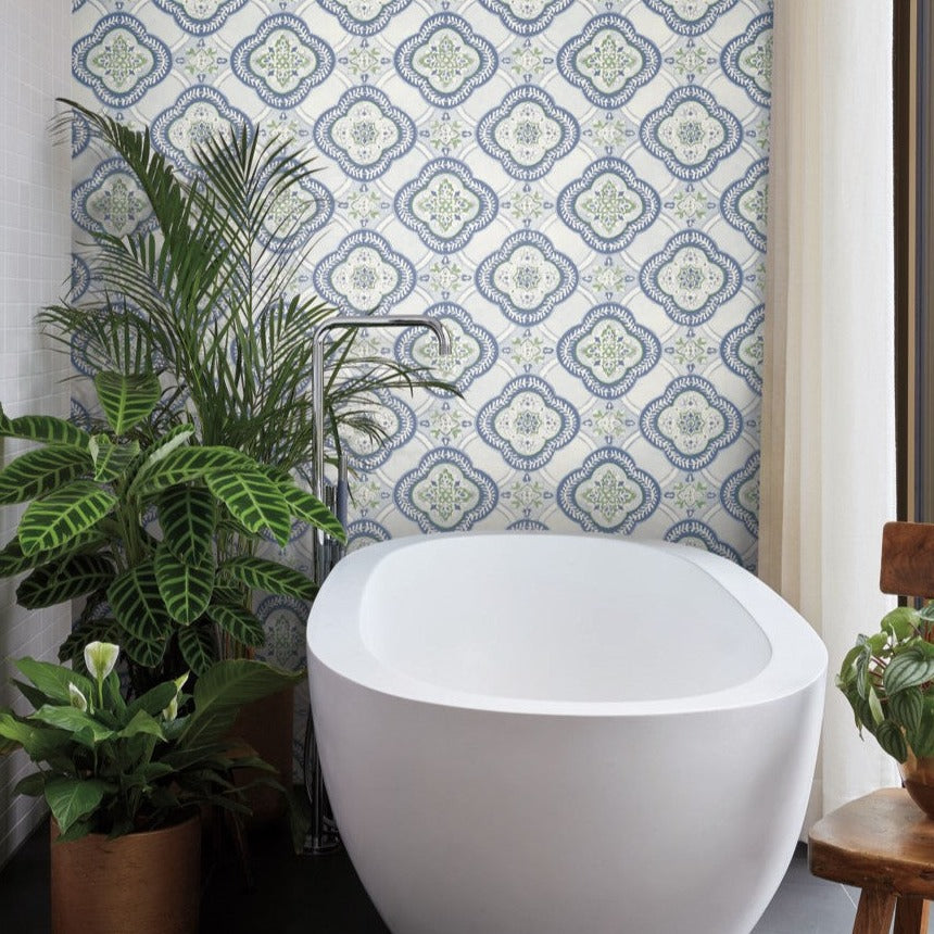 A modern bathroom exudes botanical elegance, featuring a white freestanding bathtub in front of a wall adorned with Garden Trellis Cobalt Wallpaper Blue (60 Sq.Ft.) by York Wallcoverings. To the left of the tub are tall green plants, while a sheer curtain hangs to the right. A wooden chair with a potted plant sits nearby, enhancing the serene ambiance.