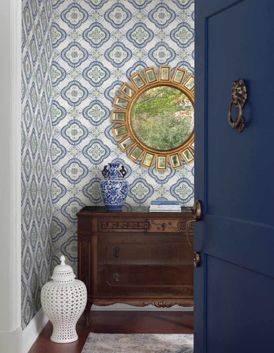 A quaint hallway features an ornate wooden sideboard, a blue and white porcelain vase, and a gold-framed round mirror on York Wallcoverings' Garden Trellis Cobalt Wallpaper Blue (60 Sq.Ft.). A large white ceramic vase sits on the wooden floor next to a blue door with a brass knocker.