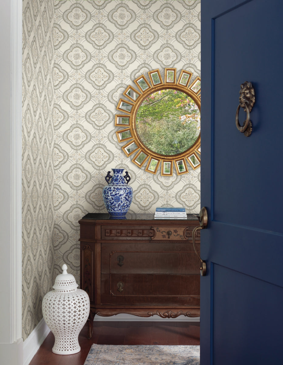 An ornate, circular mirror with a gold frame hangs on a York Wallcoverings Garden Trellis Onyx Wallpaper Black, Grey (60 Sq.Ft.) wall above a dark wooden dresser. The dresser holds a blue and white porcelain vase. A white decorative vase stands on the floor. A dark blue door is partially open, adding to the room's botanical elegance.