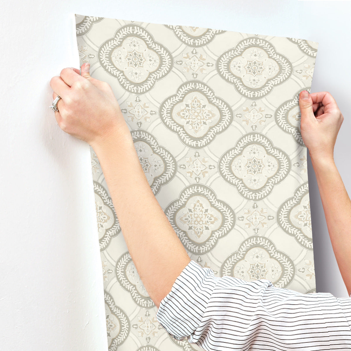 Person in a striped shirt applying York Wallcoverings Garden Trellis Rain Wallpaper Blue, Grey (60 Sq.Ft.) with a gray and white pattern on a smooth, white wall. The wallpaper features repeating intricate designs with floral and geometric motifs. The person's hands are holding the corners to ensure easy installation and removal.