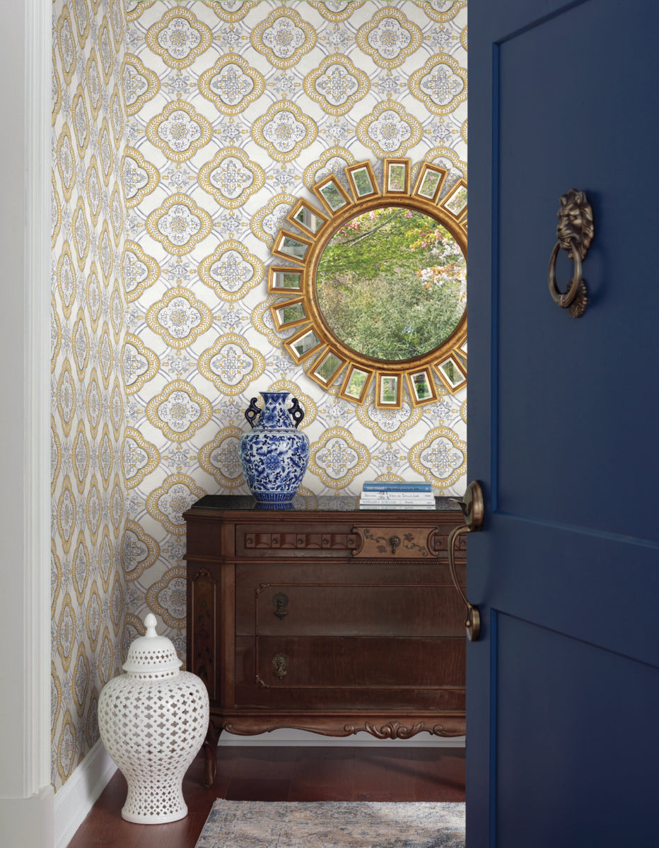 An ornate hallway featuring York Wallcoverings Garden Trellis Onyx Wallpaper Black, Grey (60 Sq.Ft.) and a wooden console with decorative drawers exudes botanical elegance. A round mirror with a gold frame hangs above the console. A blue-and-white vase and a white ceramic stool add accents, viewed through a partially open blue door.