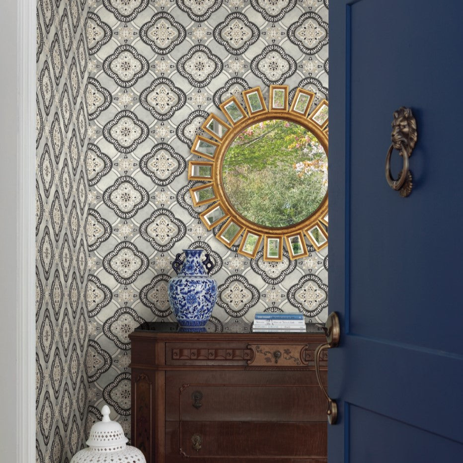 A room with botanical elegance showcases a patterned wallpaper in blue and grey, featuring the Garden Trellis Rain Wallpaper Blue, Grey (60 Sq.Ft.) by York Wallcoverings. On the wall, there's a circular gold-framed mirror. Below the mirror, there's a wooden dresser with a blue and white vase and a few books. A partially open blue door with a metallic knocker is on the right side.