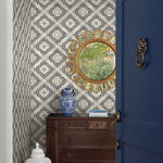 A room with botanical elegance showcases a patterned wallpaper in blue and grey, featuring the Garden Trellis Rain Wallpaper Blue, Grey (60 Sq.Ft.) by York Wallcoverings. On the wall, there's a circular gold-framed mirror. Below the mirror, there's a wooden dresser with a blue and white vase and a few books. A partially open blue door with a metallic knocker is on the right side.