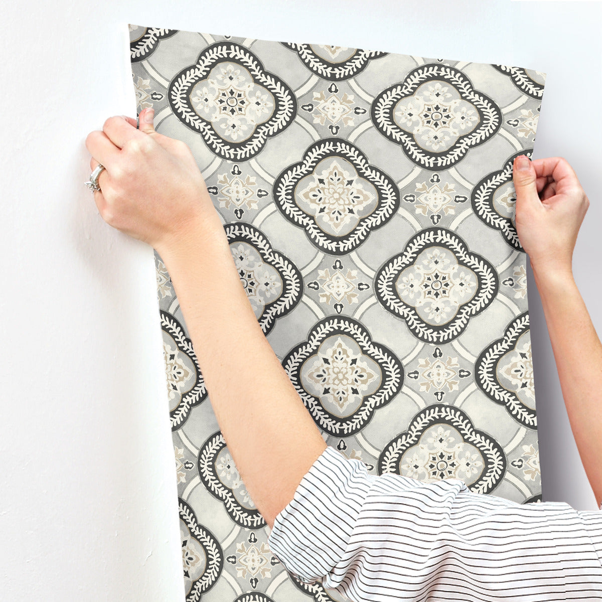 A person is hanging a sheet of York Wallcoverings Garden Trellis Rain Wallpaper Blue, Grey (60 Sq.Ft.), showcasing an ornate, symmetrical pattern in light and dark shades of gray, black, and white. The hands are positioned to smooth out the wallpaper with easy installation and removal, while wearing a white and black striped shirt.