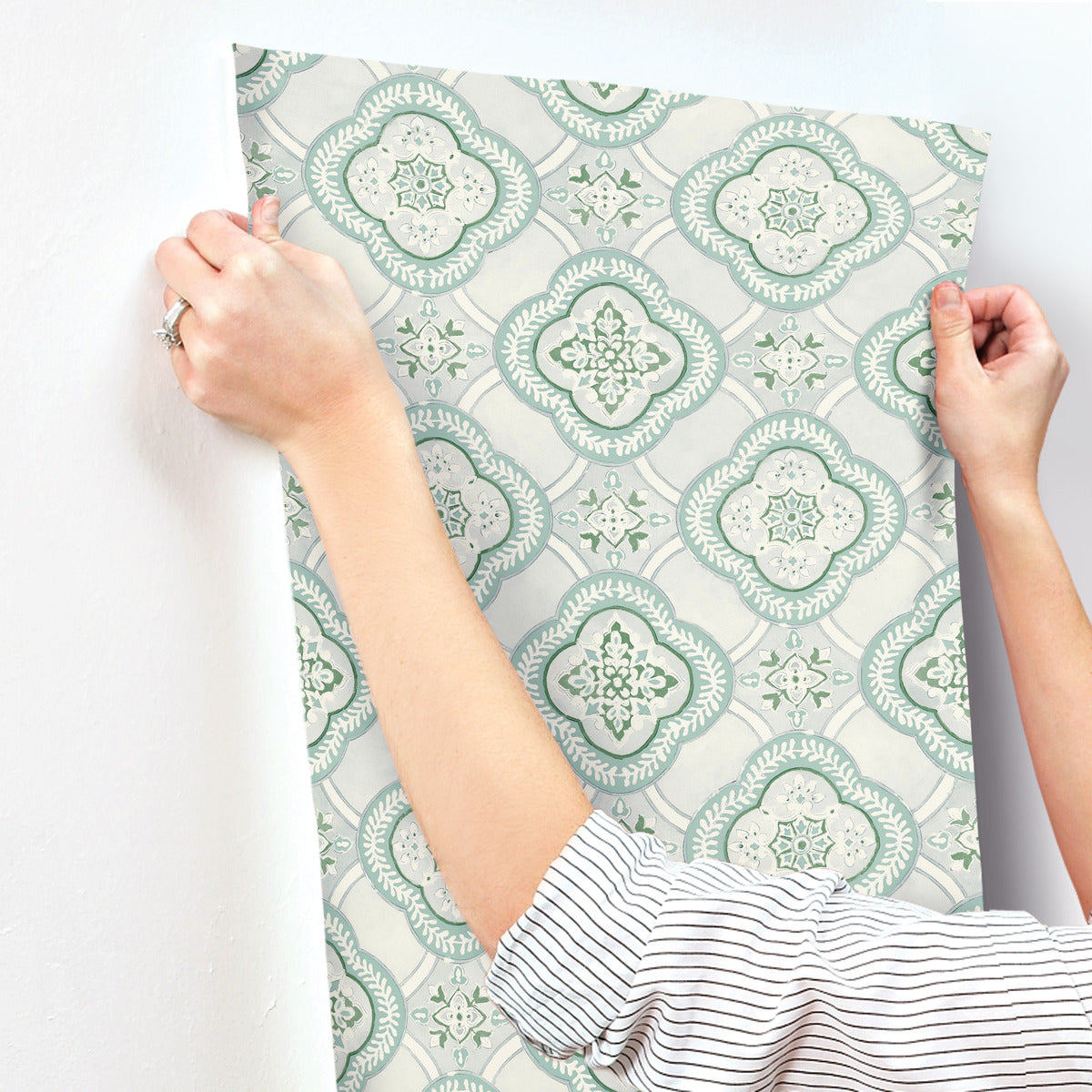 A person with a striped shirt is applying York Wallcoverings Garden Trellis Rain Wallpaper Blue, Grey (60 Sq.Ft.), featuring light blue and green patterns, to a white wall. The wallpaper design consists of intricate, symmetrical floral motifs. The person's hands are visible, smoothing out the York Wallcoverings Garden Trellis Rain Wallpaper Blue, Grey (60 Sq.Ft.) against the surface.