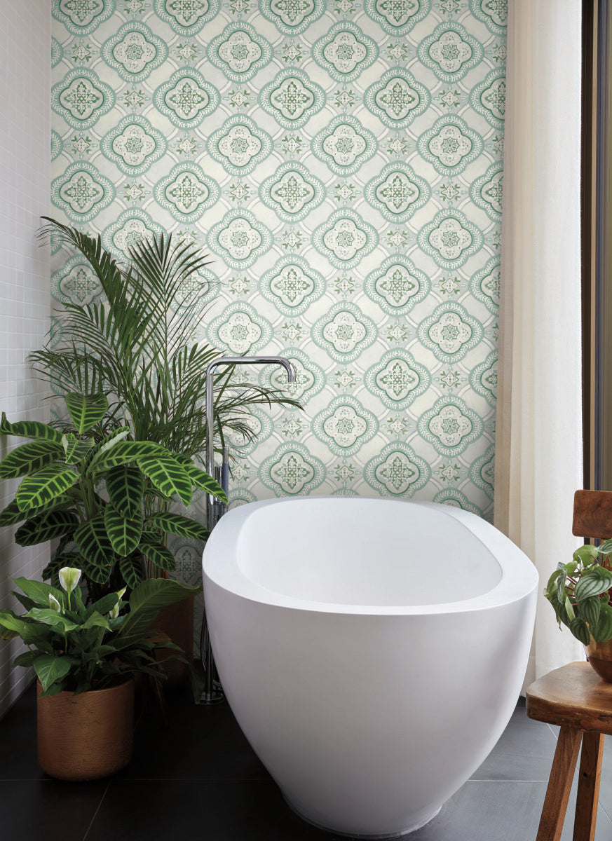 A modern bathroom with a large white freestanding bathtub against a botanical elegance of vintage-style Garden Trellis Cobalt Wallpaper Blue (60 Sq.Ft.) by York Wallcoverings. On the left are potted plants and a towel rack. Sunlight streams in from a window on the right, casting a warm light on a wooden chair and more plants.