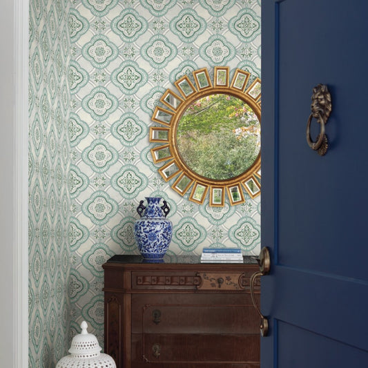 An ornate hallway features a wooden chest with intricate carvings, topped with a blue and white porcelain vase and stacked books. Above the chest hangs a round gold-framed mirror reflecting outdoor greenery. The wallpaper, presenting botanical elegance, is the Garden Trellis Rain Wallpaper Blue, Grey (60 Sq.Ft.) by York Wallcoverings.