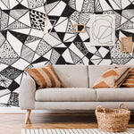 A modern living room featuring a gray sofa with patterned cushions, a wicker basket, and a striking Decor2Go Geometric Black & White Wallpaper Mural.