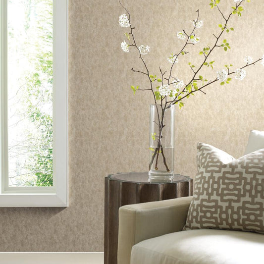 A minimalist living room features a beige sofa, a patterned cushion, and a rustic wooden side table topped with a clear vase of blooming white branches. Sunlight filters through, softly illuminating York Wallcoverings' Gold on Silver Shimmering Patina Wallpaper with its metallic stipples.