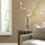 A cozy living room showcases a light beige sofa, patterned cushion, and wooden side table. A tall glass vase with white blossom branches enhances the scene. Soft natural light from a large window highlights York Wallcoverings' Gold on Taupe Shimmering Patina Wallpaper, adding elegance.