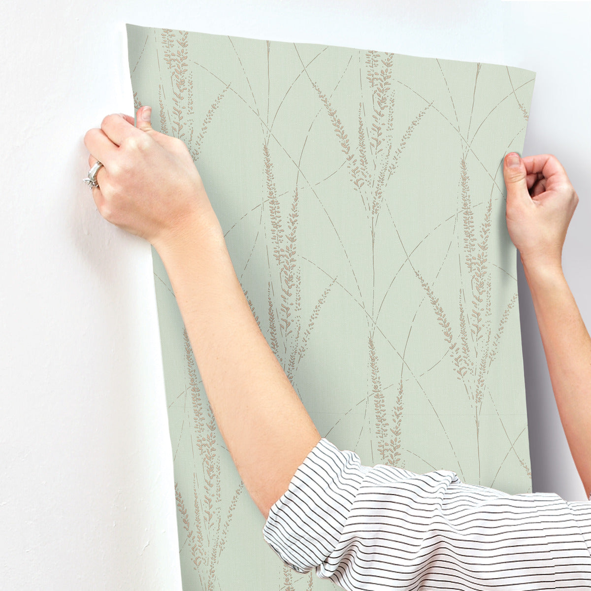 A person in a striped shirt is holding and applying York Wallcoverings' Graceful Willow Lagoon Wallpaper (60 SqFt), which features a light green botanical damask design with a beige pattern, to a wall.