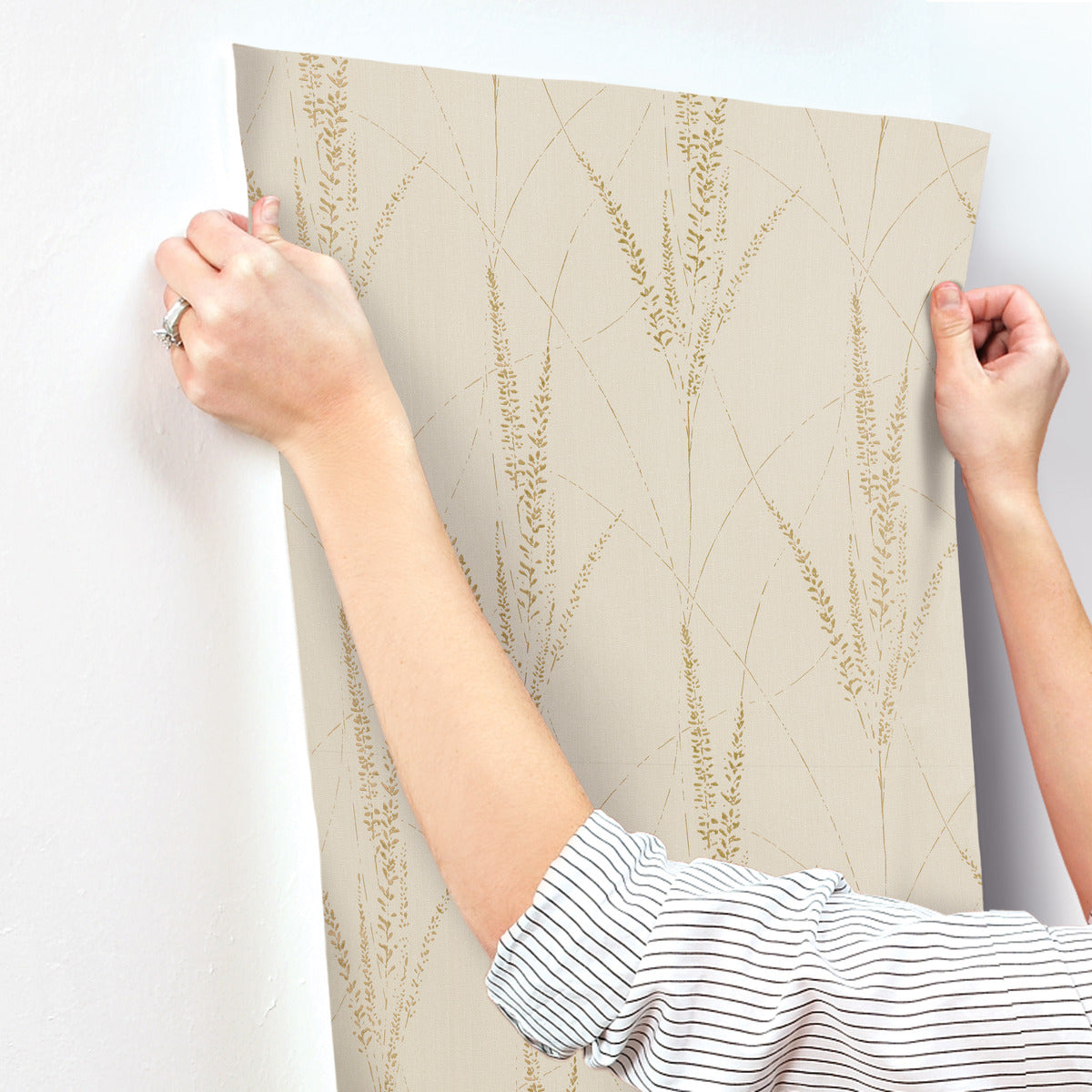 A person with a striped sleeve smooths York Wallcoverings' Graceful Willow Pale Straw Wallpaper (60 SqFt), featuring a subtle gold botanical pattern, onto a white wall.