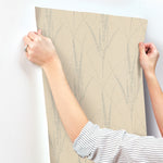 A person with a striped sleeve applies York Wallcoverings' Graceful Willow Putty Wallpaper (60 SqFt), which features a subtle light gray botanical design, to a white wall. Only their hands and arms are visible.