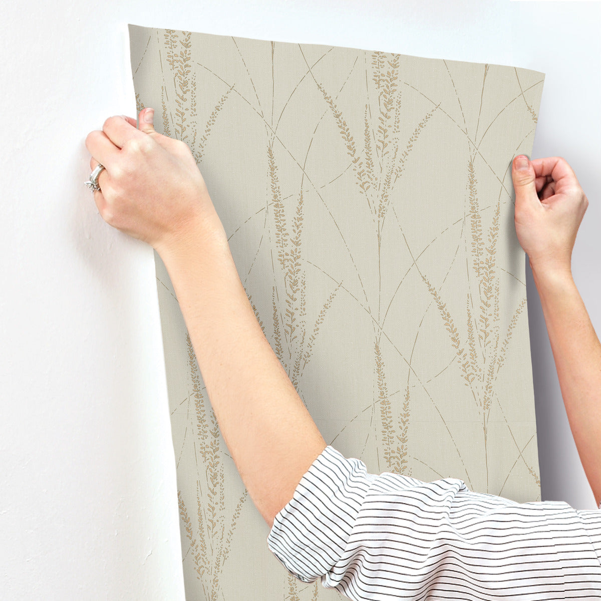 A person, wearing a striped shirt and a ring, aligns York Wallcoverings Graceful Willow Stone Wallpaper (60 SqFt) with its subtle wheat pattern against a white wall. Only their hands and forearms are visible.