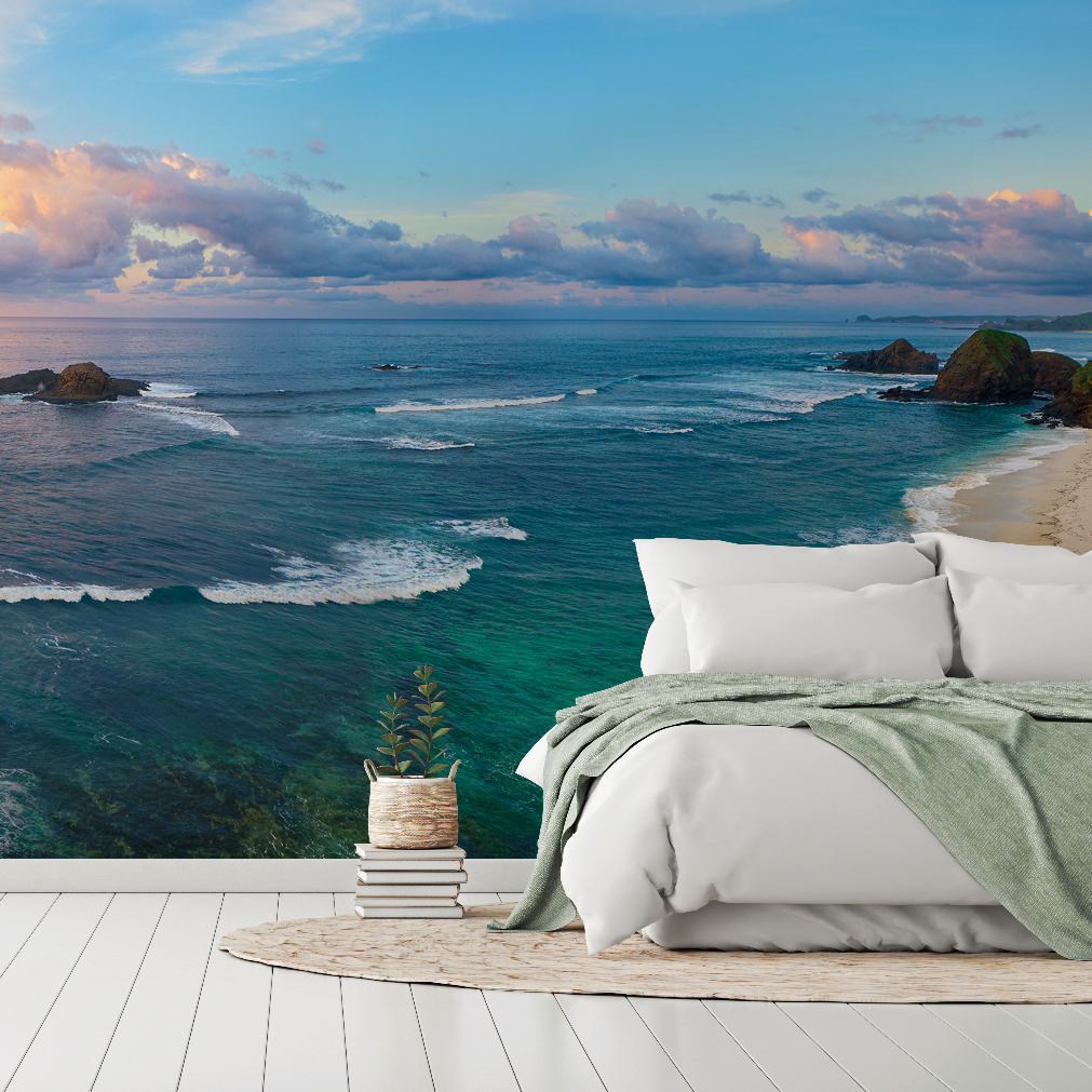 A serene bedroom setup with a bed featuring white and green bedding, situated on a wooden deck overlooking a Decor2Go Wallpaper Mural Great Blue Sea Wallpaper Mural, incorporating custom sizing to match the expansive view of clear skies and distant rocks.