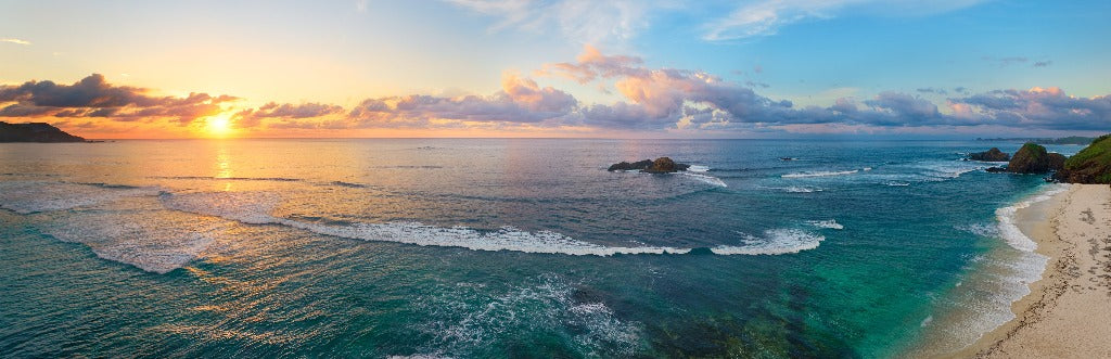 Panoramic view of a serene beach at sunrise, featuring gentle waves, a clear sky with scattered clouds, and distant rocky outcrops in the ocean Decor2Go Wallpaper Mural.