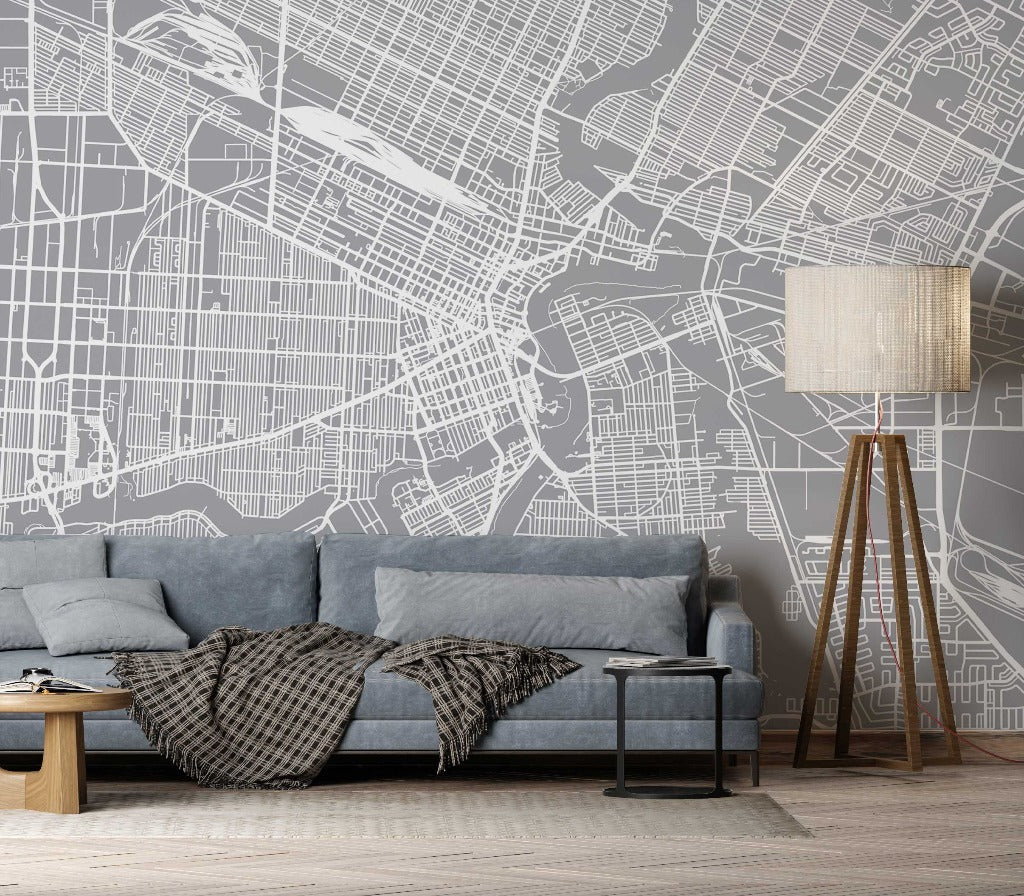 A stylish living room featuring a gray sofa with a plaid throw against a detailed Decor2Go Wallpaper Mural. A wooden tripod floor lamp and a small round table with books complete the scene.