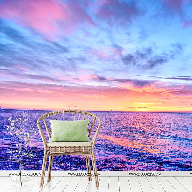 A solitary wicker chair with a green cushion placed on a seaside deck during a vibrant sunset, overlooking a calm ocean stretching to the horizon under a colorful sky featuring the Decor2Go Heaven on Earth Wallpaper Mural.