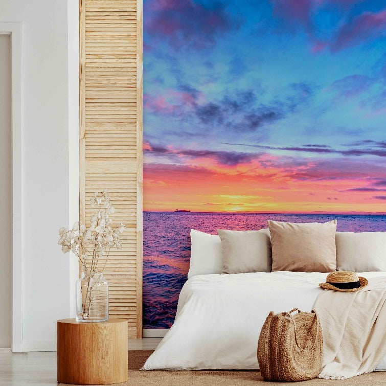 A modern bedroom with an open sliding door revealing a vivid sunset over the ocean. The room features a white bed with pillows, a wooden stool with a plant, and the Decor2Go Wallpaper Mural on the feature wall depicting Heaven on Earth.