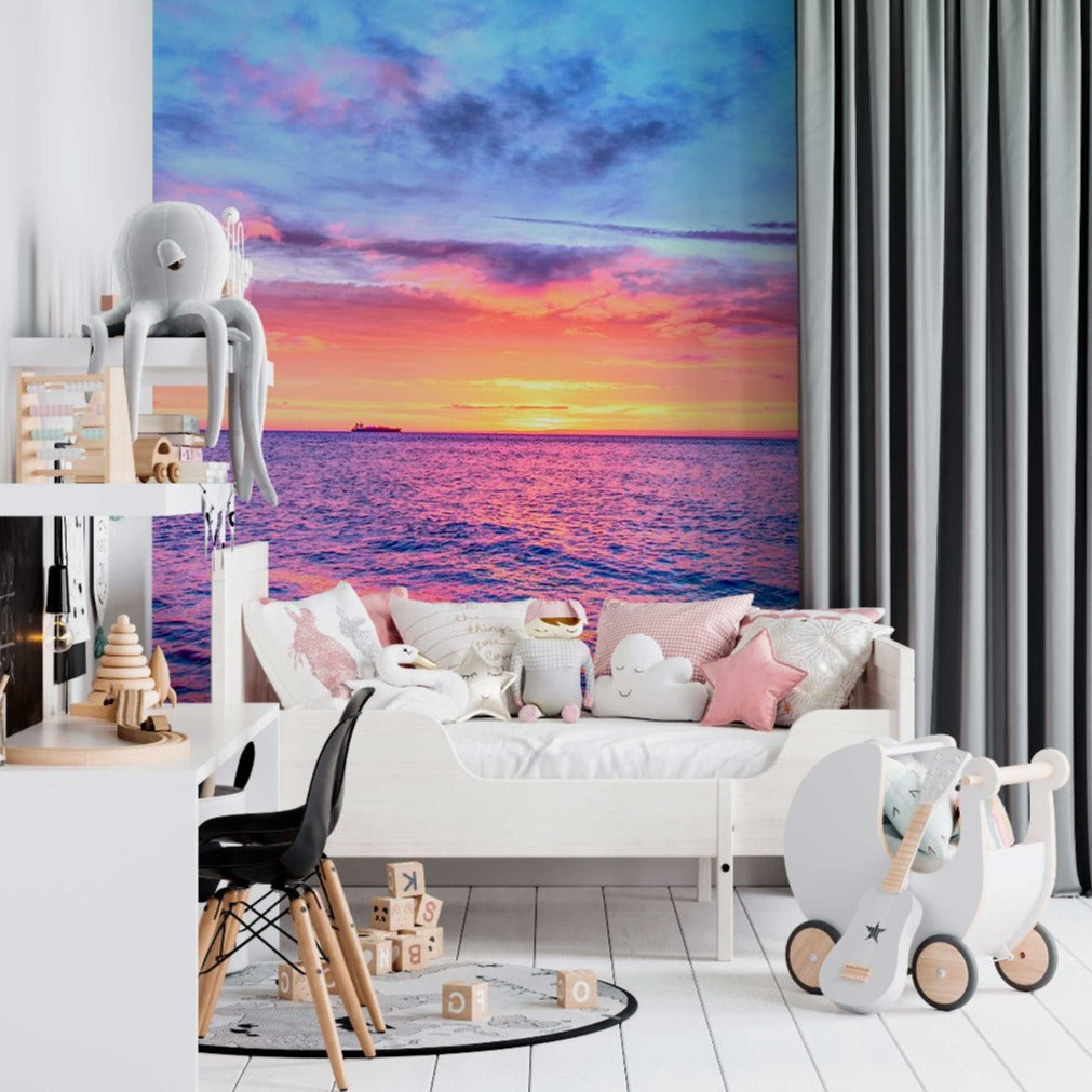 A colorful nursery room with a maritime theme, featuring a Decor2Go Wallpaper Mural of a vibrant ocean sunset. A white couch, stuffed toys, and a baby rocker are visible.