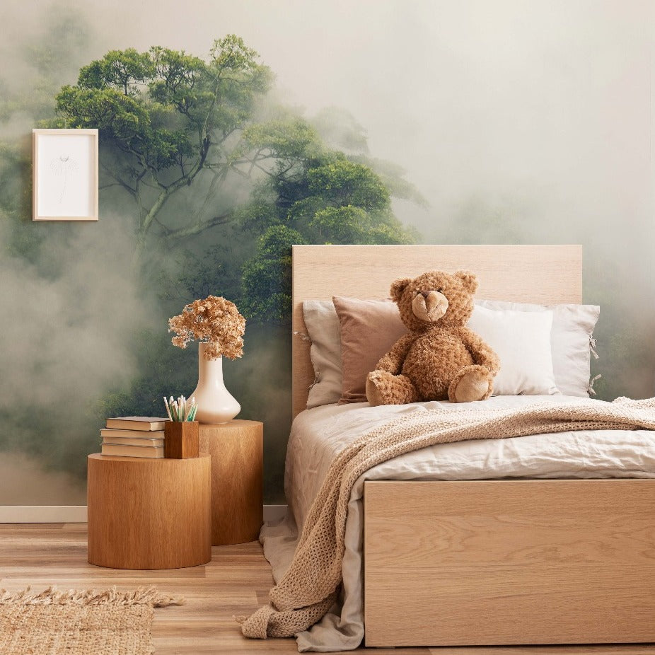A cozy bedroom featuring a wooden bed with beige and gray linens, a teddy bear sitting on the bed, and a round wooden bedside table with books and a vase of flowers. The wall has Decor2Go Wallpaper Mural's Hidden Tree Wallpaper Mural.