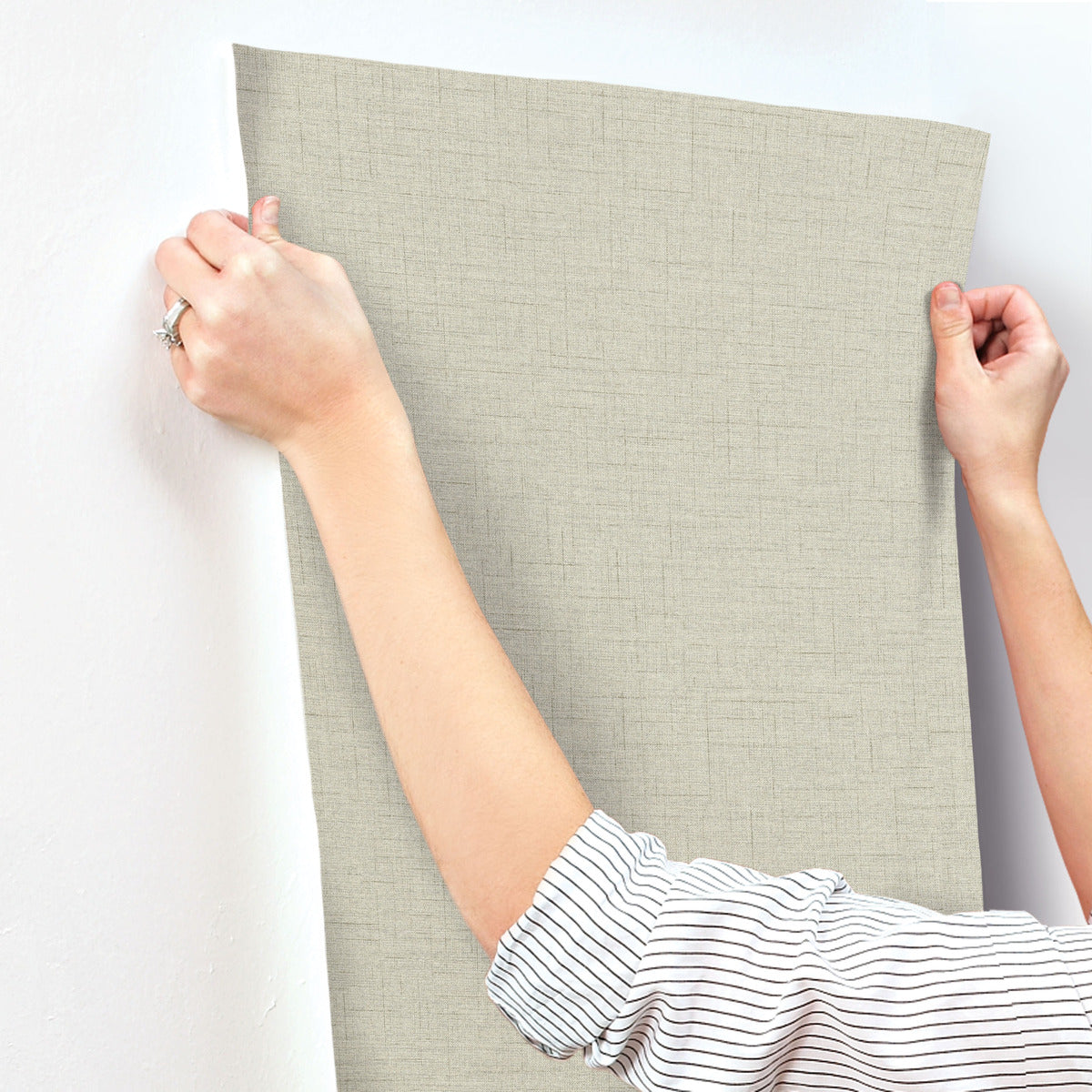A person in a striped shirt is carefully applying York Wallcoverings Hillswick Tweed Fog Wallpaper (60 SqFt), a textured non-woven beige design, to a white wall while holding it by the top corners.