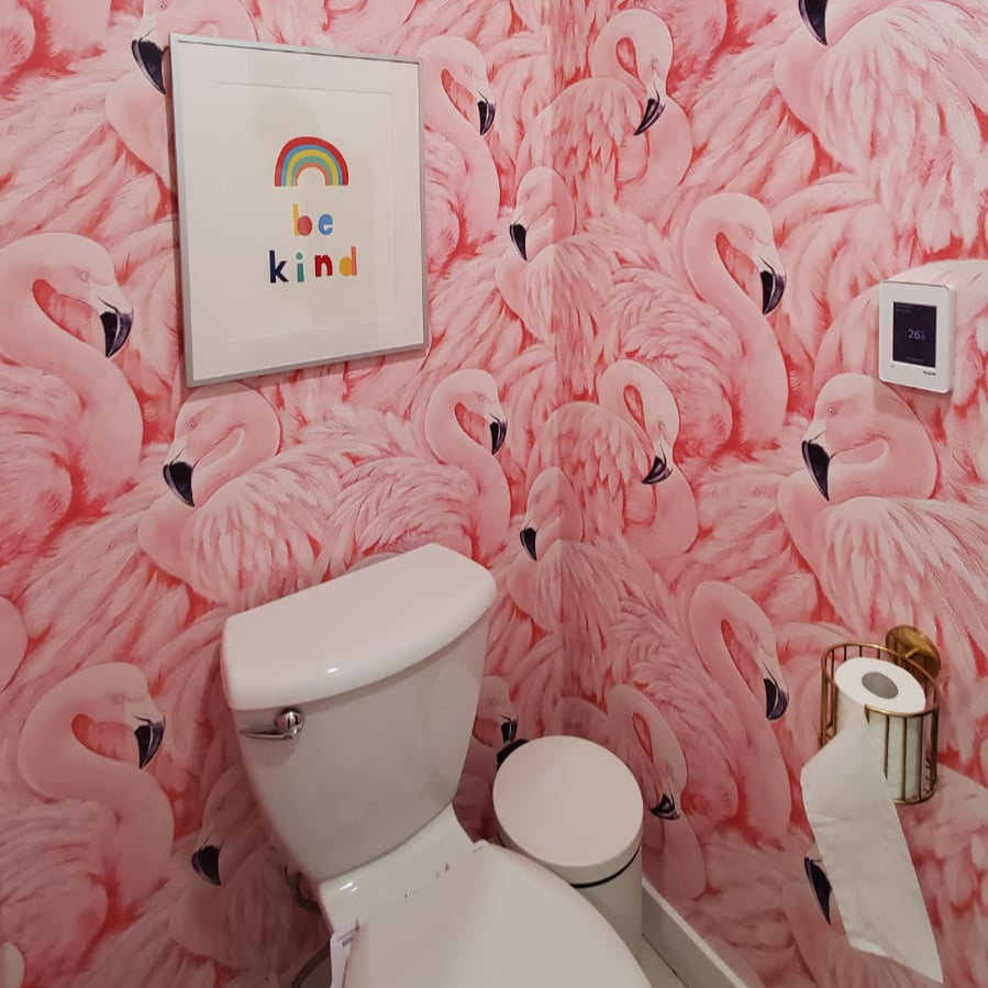 A small bathroom with Decor2Go Wallpaper Mural's Pink Flamingos Mural exudes a Tropical Escape vibe. A framed rainbow "be kind" print hangs above the white toilet, adding charm. A toilet paper holder on the right wall completes the Coastal Charm.