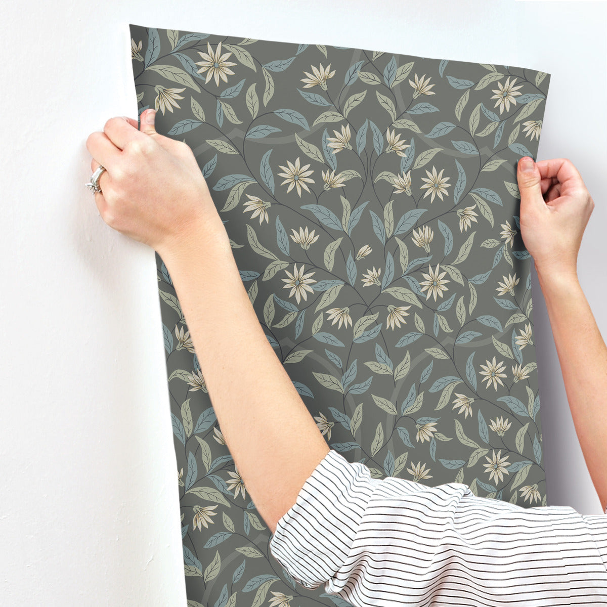 A person wearing a striped long sleeve shirt and a ring on one hand is hanging York Wallcoverings Jasmine Charcoal Wallpaper Grey, Beige (60 Sq.Ft.) with a dark background and light-colored botanical elegance. The floral pattern features leaves and flowers, creating a nature-inspired design that highlights the SureStrip Substrate.