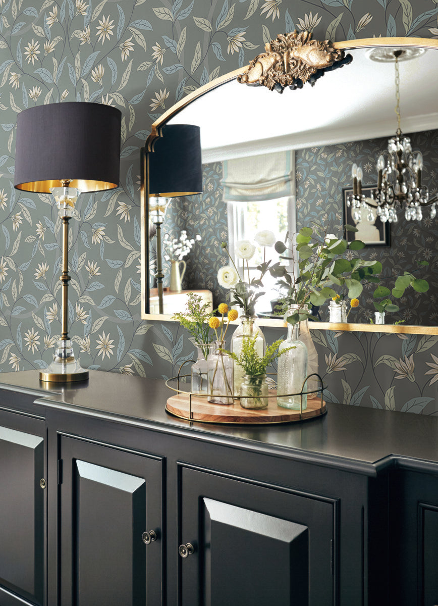 A decorated room with a large mirror above a black sideboard. The wall is covered in Jasmine Charcoal Wallpaper Grey, Beige (60 Sq.Ft.) by York Wallcoverings with a floral pattern. On the sideboard, a tray holds various glass bottles with small plants, and a black and gold lamp stands beside them. A chandelier is reflected in the mirror.