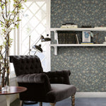 A cozy reading nook with a dark tufted armchair beside a small round wooden table adorned with greenery in clear vases. The wall behind features York Wallcoverings Jasmine Charcoal Wallpaper Grey, Beige (60 Sq.Ft.), and two white shelves display books and an art piece. Soft natural light streams in from the window.