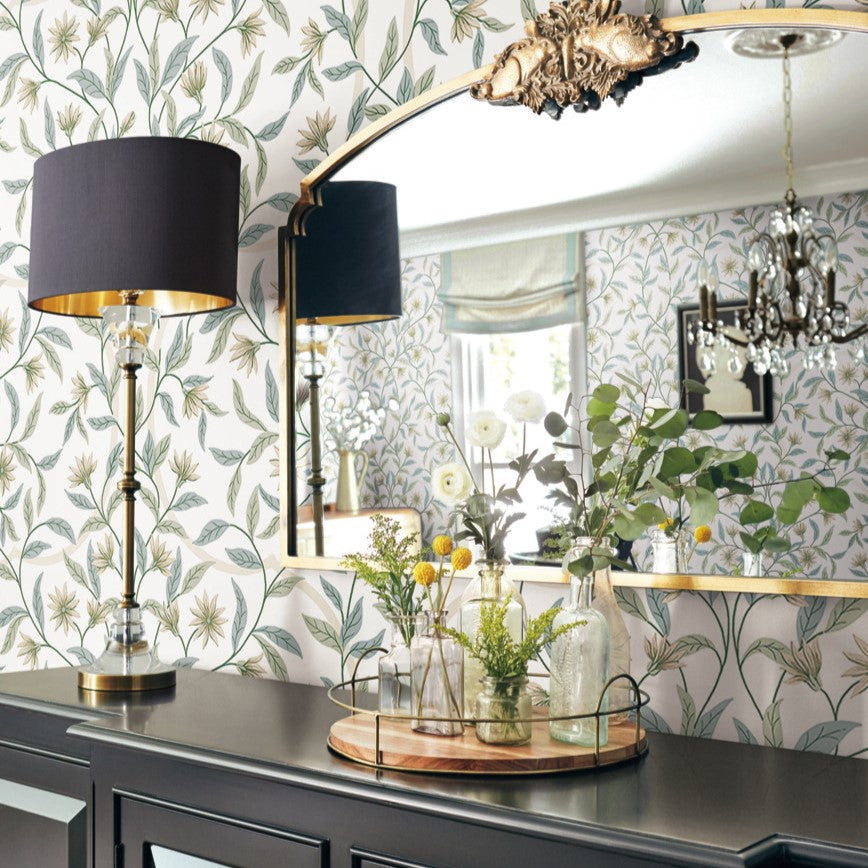 A room with Jasmine Cornflower Wallpaper Beige, Blue (60 Sq.Ft.) by York Wallcoverings featuring a large mirror with an ornate gold frame. In front of the mirror is a dark-colored sideboard with a decorative tray holding various vases and flowers. To the left stands a modern black and gold lamp, adding to the space's botanical elegance.