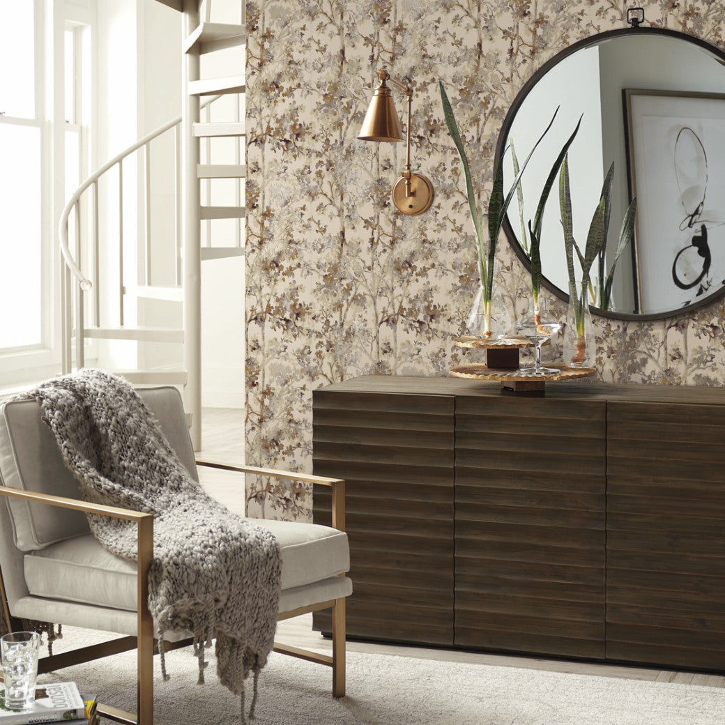 A cozy living room features a beige armchair and knitted throw. On the right, a dark wooden dresser is topped with items from the Modern Metals Collection, including a copper vase. The Khaki & Multi Shimmering Foliage Wallpaper by York Wallcoverings hosts a round mirror, with a spiral staircase in view.