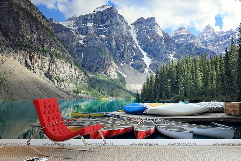 A serene mountain lake setting with canoes lined up on a rustic wooden dock, surrounded by lush green forests and rugged peaks, featuring a red lounge chair facing the water with Decor2Go Wallpaper Mural's Lakeside Escape Wallpaper Mural.