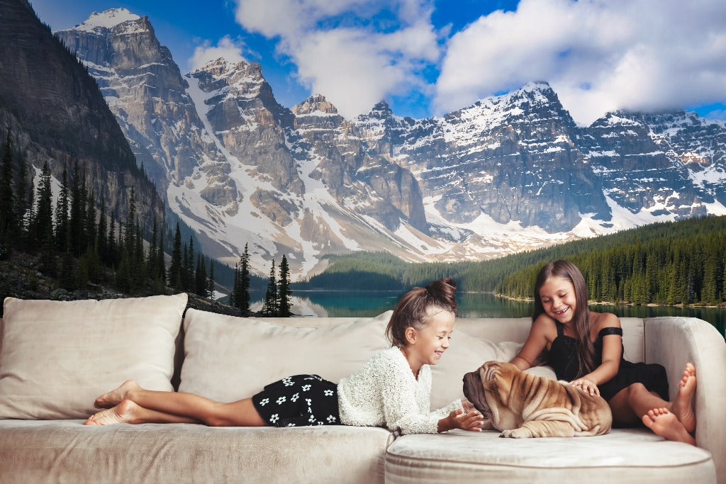 Two young girls and a dog playfully interact on a sofa in front of a feature wall with the Decor2Go Wallpaper Mural depicting majestic mountains and a lush forest around a lake.