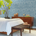 A stylish bedroom showcases a bed adorned with white and light gray blankets. A wicker bench holds a tan hat, while the wooden dresser is arranged with books and vases. The nature-inspired design is accentuated by a large leafy plant set against vibrant York Wallcoverings' Laurel Damask Wallpaper in blue.