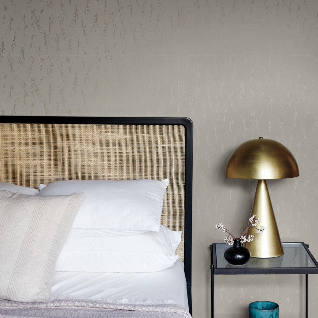 A modern bedroom showcases a wicker-backed bed with white pillows and a lavender throw. A sleek nightstand holds a gold lamp, complementing a small branch in a vase. The standout feature is the York Wallcoverings' Light Grey & Gold Sprigs Wallpaper, providing a textured elegance to the wall.