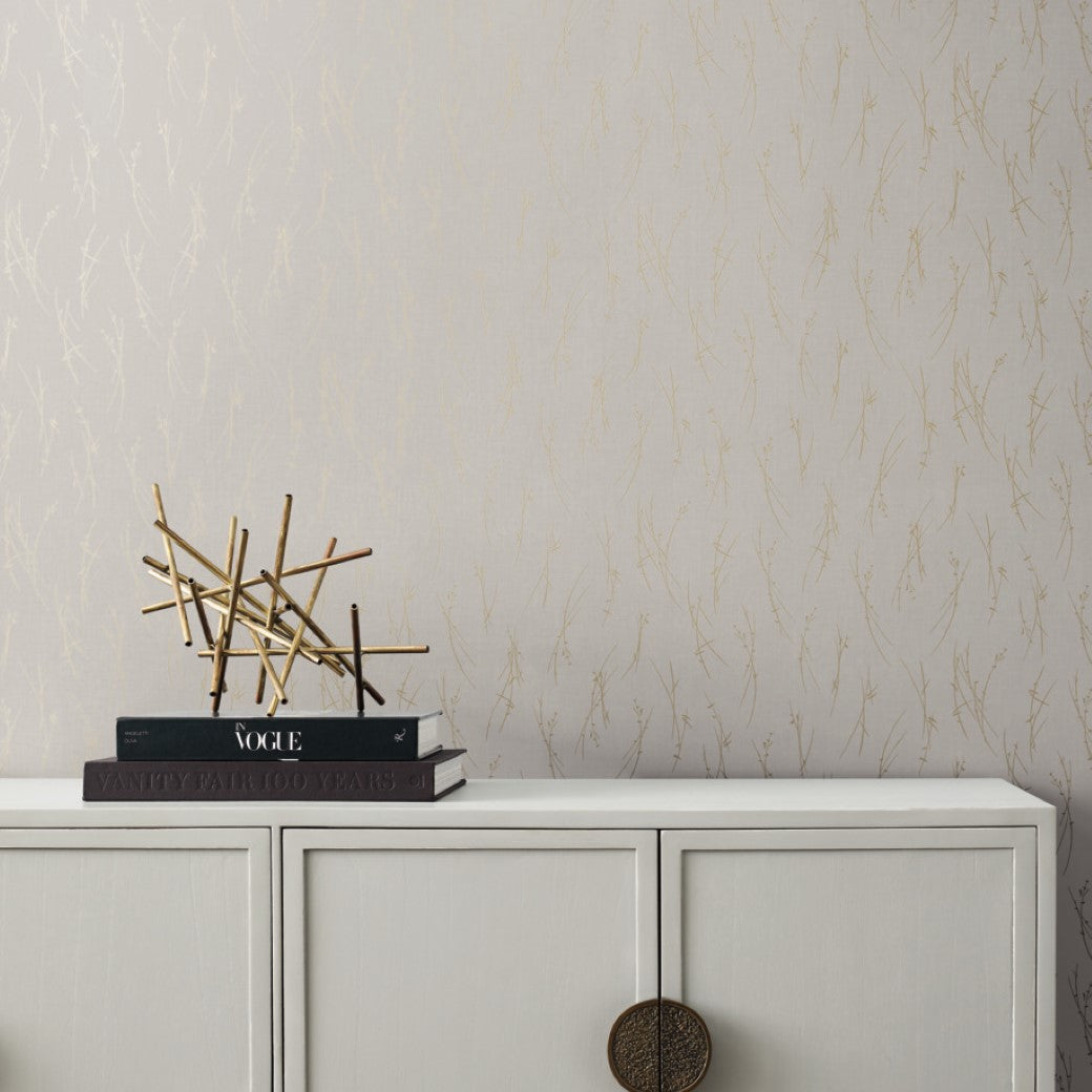 A modern interior scene showcases a minimalist white cabinet with round bronze handles. Art books stack beside a golden geometric sculpture, enhancing the ambiance. The Light Grey & Gold Sprigs Wallpaper by York Wallcoverings ties it together with its elegant light beige and gold patterns.